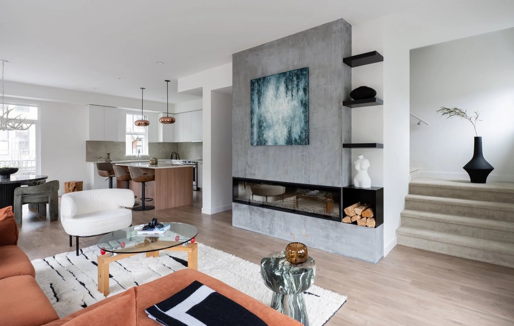 Britannia Beach House — Open living area with a concrete fireplace, rust sofa and steps to an adjoining space