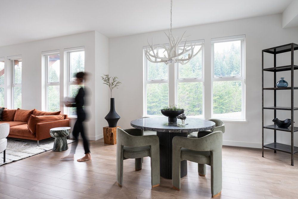 Britannia Beach House — Round dining table with green chairs, rust sofa and a figure moving past tall windows